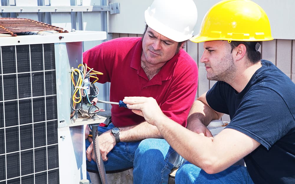 Two HVAC technicians working on the wiring for an AC unit.