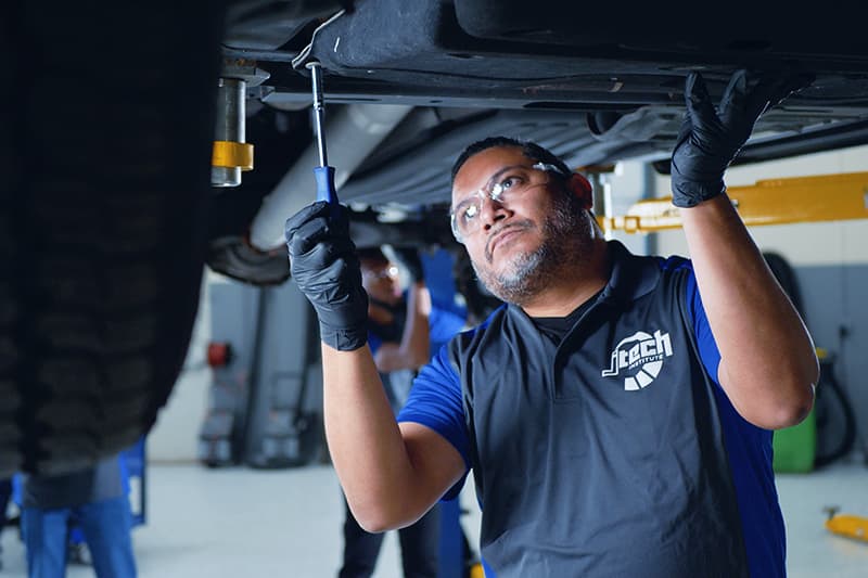 An older J-Tech Automotive Technology student unscrewing a cover panel underneath a car on a lift.