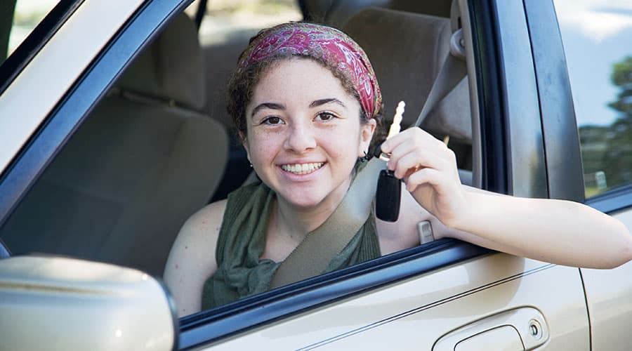 A teenage girl getting into her first car, holding the key and smiling.